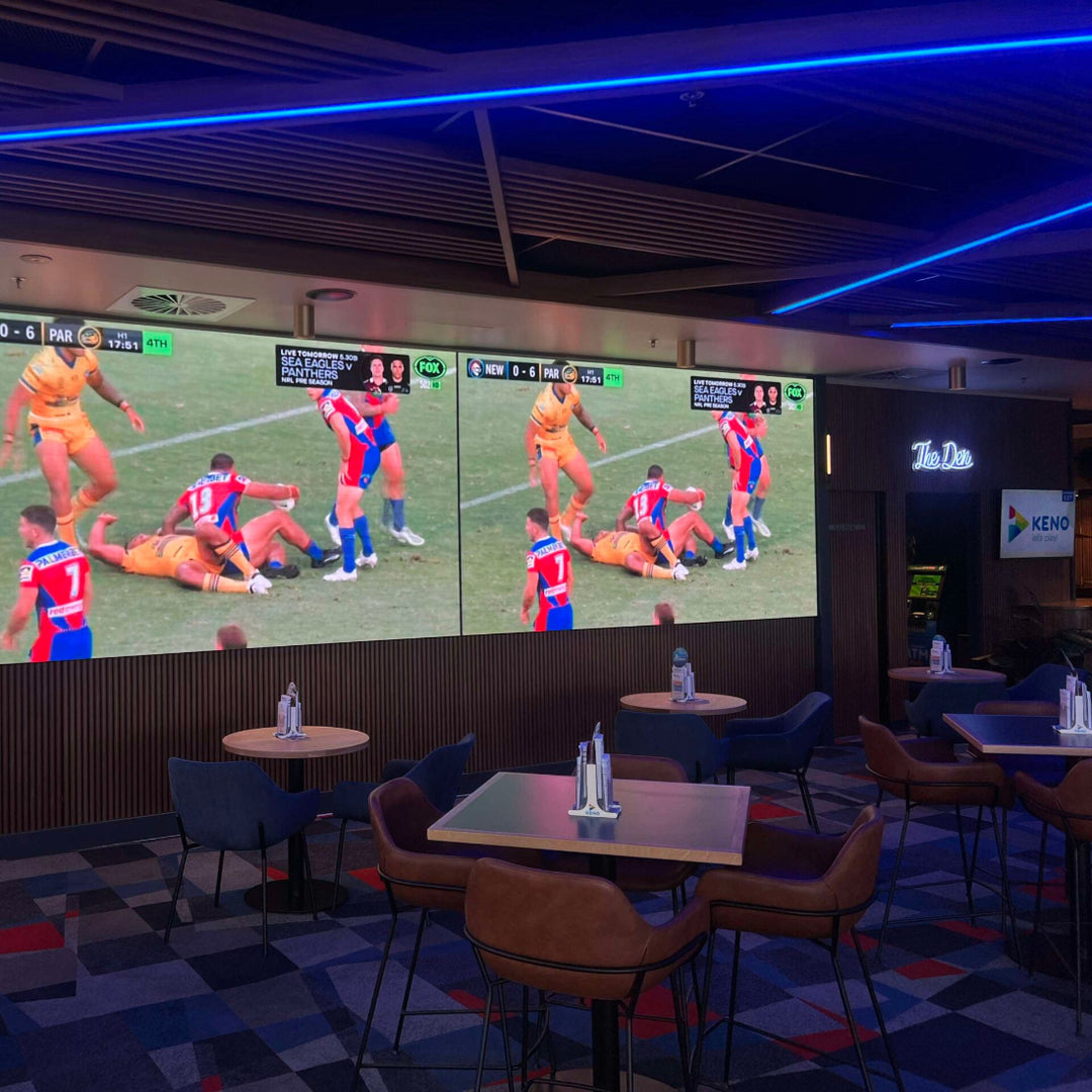 Bar or sports bar with a large screen displaying a rugby match, tables, and chairs. Overhead RGB linear surface mount lighting shines down to create a ambient experience 